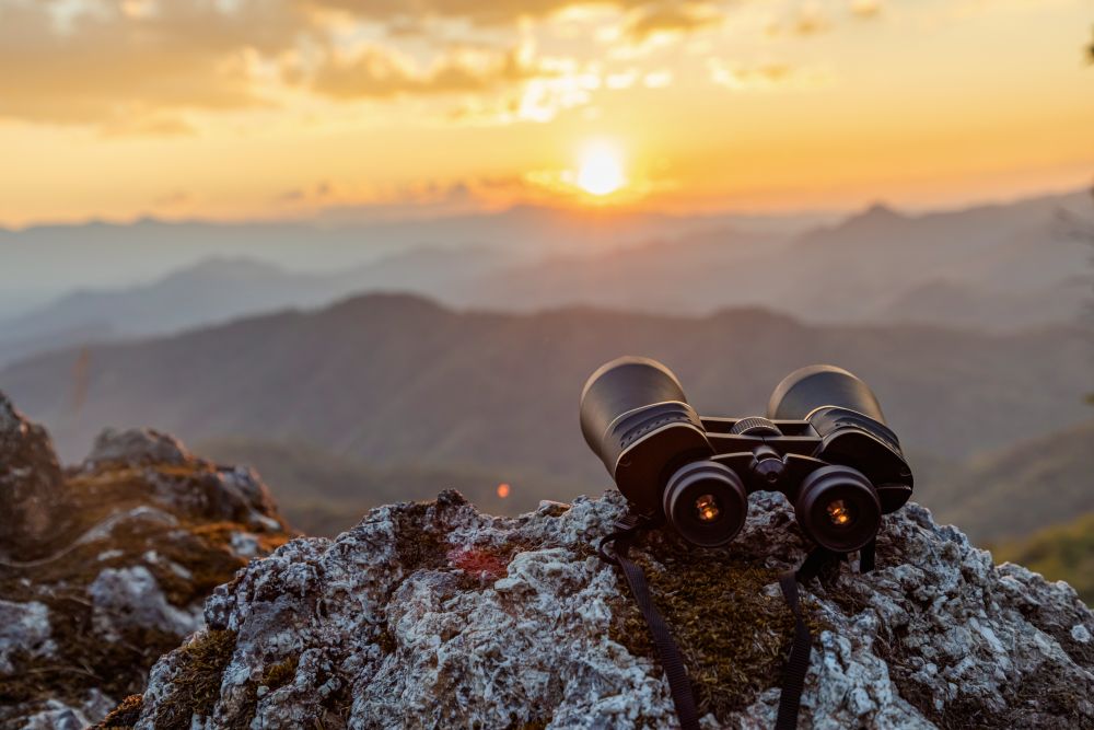 Ein Fernglas liegt bereit zur Vogelbeobachtung mit einer Berglandschaft bei Sonnenuntergang im Hintergrund. 